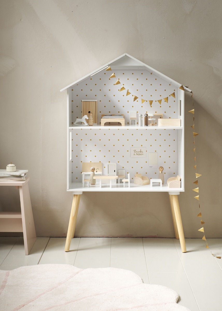Maison de poupée blanche sur pieds en bois, aménagée avec des meubles et figurines en bois, décorée de guirlandes et d’un papier peint à pois dorés, dans une chambre d’enfant aux tons doux.